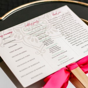 May include: A white wedding ceremony program with a pink ribbon and wooden handle. The program lists the order of the ceremony and the names of the wedding party members. The text at the bottom reads "All our love, Sarah & William".