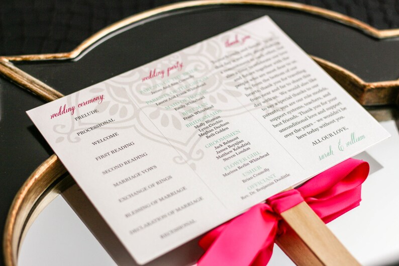 May include: A white wedding ceremony program with a pink ribbon and wooden handle. The program lists the order of the ceremony and the names of the wedding party members. The text at the bottom reads "All our love, Sarah & William".