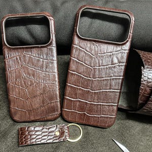May include: Two brown leather phone cases with a crocodile skin texture are displayed. A matching keychain with a gold ring is also present. The cases have a rectangular shape with a camera cutout. The leather is a rich, dark brown color.