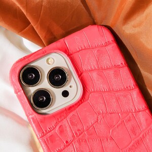 Puede incluir: Una funda de teléfono de color rosa brillante con textura de piel de cocodrilo. La funda tiene una protuberancia de cámara elevada con tres lentes.
