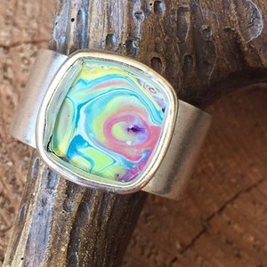 May include: A silver ring with a colorful, abstract design set in a square-shaped resin. The resin is a swirl of pink, blue, green, and yellow.