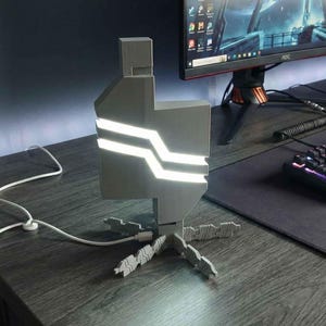 May include: A gray and white LED desk lamp with a unique geometric design. The lamp has two horizontal white light strips and a white power cord. The lamp is on a dark wood desk with a computer monitor, keyboard, and mouse.