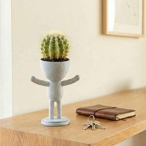 May include: A grey, human-shaped planter holding a green cactus. The planter has outstretched arms and a round base. A brown leather notebook and keys sit on a wooden surface.