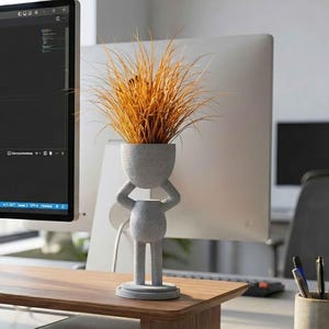 May include: A gray concrete planter shaped like a person holding its head, filled with dried orange grass. The planter has a round base and is on a wooden desk. The planter is approximately 10 inches tall.