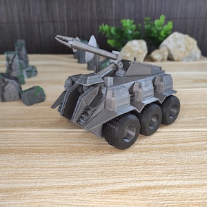 May include: A gray, detailed, futuristic-style toy vehicle with a raised, articulated weapon and four wheels. The vehicle is on a wooden surface.
