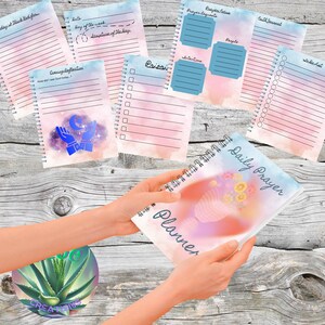May include: A Daily Prayer Planner with a floral design is held in hands. The planner has a spiral binding and includes pages for daily reflections, priorities, and prayer requests. Other pages include a faith journal and a to-do list.