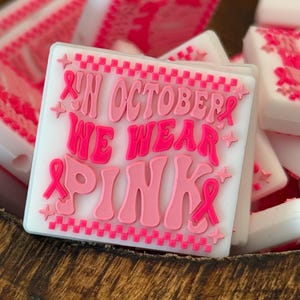May include: A collection of white square-shaped items with pink text that reads "IN OCTOBER WE WEAR PINK". The text is in a retro font with pink ribbons and stars. The items are in a wooden bowl.