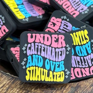 May include: Black square charm with the text "UNDER CAFFEINATED AND OVER STIMULATED" in pink, purple, blue, and yellow. The charm has a retro font and star accents. Other charms with similar text are in the background.