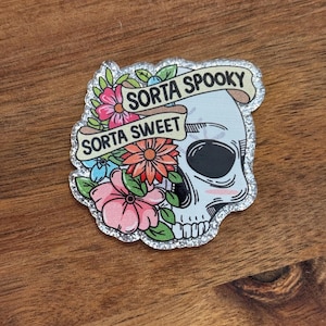 May include: A decorative sticker featuring a skull with floral accents in pink, orange, and blue. The sticker has the phrases "Sorta Spooky" and "Sorta Sweet" on banners. The edges are outlined with silver glitter.