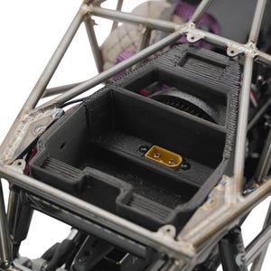 Könnte beinhalten: Nahaufnahme einer schwarzen rechteckigen Komponente mit einem gelben Stecker, eingebettet in einen silbernen Metallrahmen. Der Rahmen ist Teil eines Fahrzeugs oder einer Maschine, mit sichtbaren Schweißnähten und Halterungen. Der Hintergrund ist unscharf.