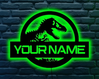 May include: A black metal sign with a green neon glow, featuring a dinosaur silhouette within a circular frame. The text "YOUR NAME" is displayed in a rectangular panel below the circle. The sign is mounted on a brick wall.