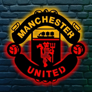 May include: A black and red Manchester United logo with the words "MANCHESTER UNITED" in a bold font. The logo features a red devil holding a trident, with a yellow and red illuminated background. Two footballs are on either side of the logo.