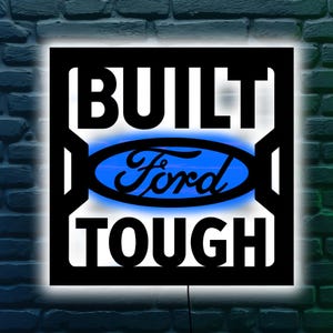 May include: Black metal sign featuring the words "BUILT TOUGH" and the Ford logo in a blue oval. The sign is illuminated with a white and blue glow, mounted on a brick wall.