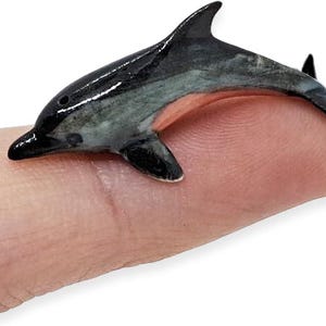 May include: A miniature figurine of a dolphin with a glossy finish, featuring a black dorsal side, a white and grey mottled midsection, and a light pink underbelly, captured in a scale that fits on the tip of a human finger for size reference.