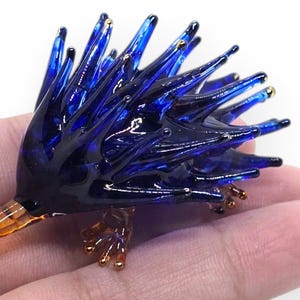May include: A handcrafted glass hedgehog figurine. The figurine features a dark blue body with spiky protrusions and amber-colored feet and snout. The glass sculpture is held in a hand against a white background.
