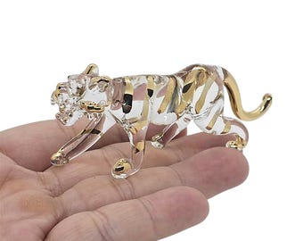 May include: A clear glass tiger figurine with gold accents. The tiger is depicted in a walking pose, with gold stripes and details on the paws, ears, and tail. The figurine is held in a hand.