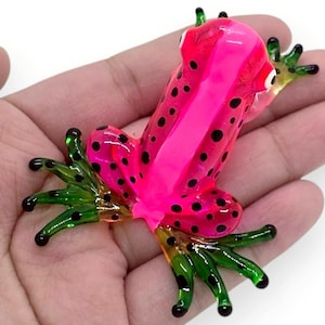 May include: A vibrant, glass frog figurine in shades of pink and green. The frog has a bright pink body with black spots and a vertical stripe. Its feet are green with black spots. A decorative item.