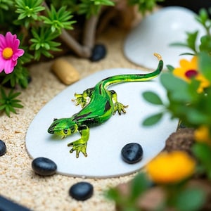 May include: A detailed glass lizard figurine in shades of green and yellow rests on a white oval. The lizard is surrounded by small black stones, sand, and miniature plants with pink and yellow flowers. The figurine has a curled tail.