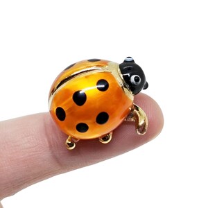 May include: A small, orange ladybug figurine with black spots and a black head. The ladybug has gold-colored legs and a gold trim around its body. The figurine is held on a finger.