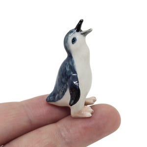 May include: A small, ceramic penguin figurine with a white body, blue-grey wings and back, and black beak. The penguin has blue eyes and is posed with its beak open. The figurine is held on a finger.