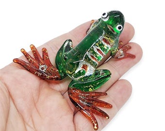 May include: A handcrafted glass frog figurine. The frog is primarily green with red-orange feet and accents. The glass is translucent, allowing light to pass through, and features decorative gold flecks. The frog's eyes are white with black pupils.