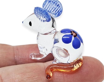 May include: A clear glass mouse figurine with blue accents. The mouse has blue ears and a blue and white floral design on its back. The tail is amber-colored. The figurine is small and detailed.