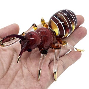 May include: A detailed glass ant sculpture with a segmented body. The ant features a dark red head and thorax, with a striped amber and white abdomen. The legs and antennae have gold accents.