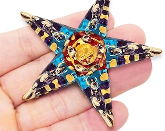 May include: A star-shaped decorative object with gold, blue, purple, and red accents. The center features a gold emblem, and the star points have a gold finish. The object is held in a hand.