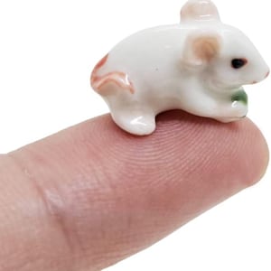 May include: A miniature white ceramic mouse figurine with pink-tinted ears and tail, featuring a small dark eye and holding a tiny green object in its front paws. The smooth, glossy surface reflects light, highlighting the rounded, compact form.