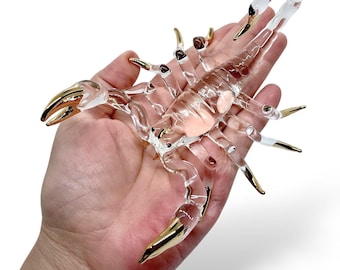 May include: A clear glass scorpion figurine with gold-colored accents on the claws and legs. The scorpion is held in a hand against a white background. The glass is transparent, allowing light to pass through.