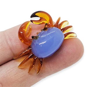 May include: A miniature glass crab figurine. It has a light blue body and amber claws and legs. The crab has small black eyes. The figurine is resting on a finger. The crab is a decorative item.