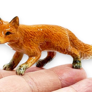 May include: A small, hand-painted ceramic fox figurine featuring a glossy finish, vibrant orange-brown fur, pointed ears, and dark-tipped paws. The fox is depicted in a standing, alert posture with its tail extended, showcasing detailed fur texture.