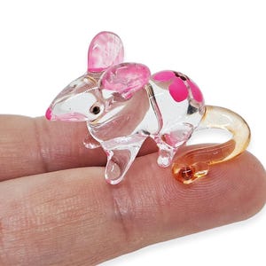 May include: A clear glass mouse figurine with pink accents on the ears and back. The mouse has a curled amber-colored tail and small black eyes. The figurine is a miniature decorative item.
