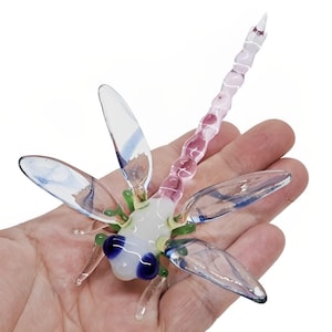 May include: A handcrafted glass dragonfly sculpture. The dragonfly features clear wings with blue accents, a white body, and a pink and white tail. The eyes are blue, and the legs are green. The sculpture is held in a hand.
