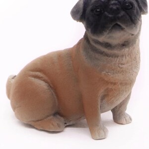 May include: A small, detailed figurine of a pug dog. The pug is depicted in a seated position with a tan body and a black face. The figurine has a smooth texture and is set against a white background.