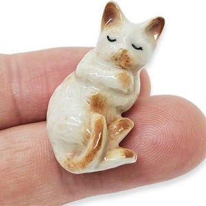 May include: A small, ceramic cat figurine in a seated position with its eyes closed. The cat is primarily white with brown markings on its ears, paws, and body. The figurine has a glossy finish and appears to be handcrafted.