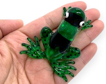 May include: A green glass frog ornament with black stripes and black spots. The frog has large eyes and outstretched limbs. The ornament is held in a hand against a white background. The frog is a decorative item.