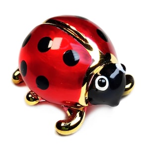 May include: A shiny red ladybird ornament with black spots and gold-coloured detailing. The ladybird has a black head with white eyes and gold feet. The ornament is a decorative item.