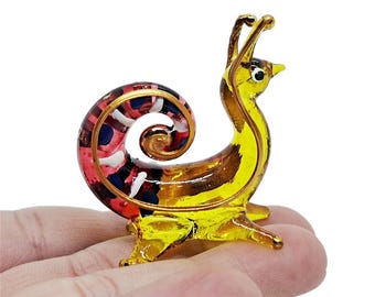 May include: A glass snail figurine with a yellow body, brown stripes, and a colorful shell. The shell features red, white, and blue accents. The figurine is held in a hand.