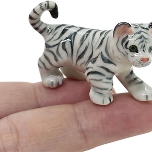 May include: A small ceramic tiger figurine. The tiger is white with black stripes, green eyes, and a pink nose. It is posed in a walking stance. The figurine is held in a person's hand.