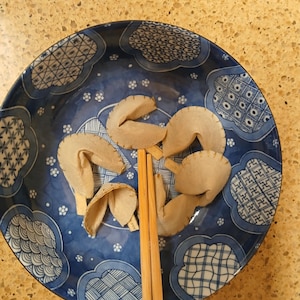 Può includere: Un piatto blu e bianco a motivi contiene diversi biscotti della fortuna beige. Due bacchette di legno sono appoggiate sul piatto. Il piatto ha un design decorativo con motivi floreali e geometrici.