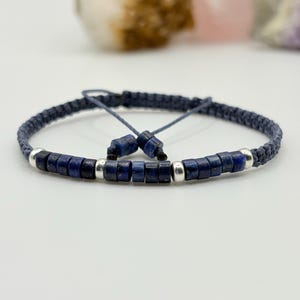Op de afbeelding: Een handgemaakte armband met donkerblauwe kralen en zilverkleurige accenten. De armband is gemaakt van een geweven, donkerblauw koord en heeft een verstelbare sluiting. De kralen zijn rechthoekig en gelijkmatig verdeeld.