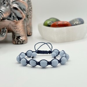 Könnte beinhalten: Ein hellblaues Perlenarmband mit einer dunkelblauen geflochtenen Kordel. Das Armband hat runde, hellblaue Perlen. Im Hintergrund eine dekorative Elefantenstatue und eine Schale mit bunten Steinen.