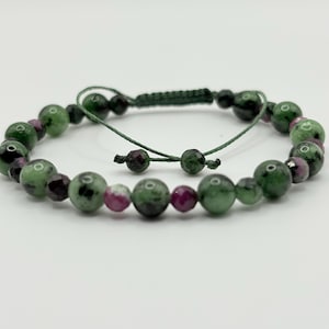 Może przedstawiać: Regulowana bransoletka z połączeniem zielonych i rubinowych koralików. Okrągłe koraliki różnią się rozmiarem i są nawleczone na ciemnozielony sznurek. Posiada zapięcie na przesuwny węzeł dla indywidualnego dopasowania. Design jest prosty i elegancki.