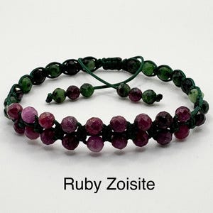 May include: A double-strand bracelet with faceted ruby zoisite beads in pink and burgundy, strung on a green cord. The bracelet has an adjustable sliding knot closure. The text "Ruby Zoisite" is displayed below the bracelet.