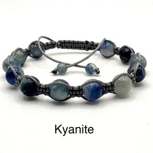 May include: A macramé bracelet featuring blue and grey kyanite beads. The adjustable bracelet has a sliding knot closure and the word "Kyanite" is printed below the bracelet.