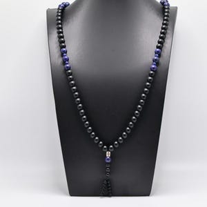 May include: A black beaded necklace with blue accents. The necklace is displayed on a black mannequin. The beads are small and round, with a few larger blue beads interspersed throughout. The necklace has a long, dangling pendant.