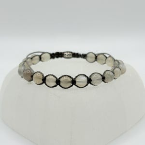 May include: A macrame bracelet featuring light gray, round beads. The beads are strung on a black cord, creating a woven pattern. The bracelet is displayed on a white, geometric surface, showcasing the jewelry's design and craftsmanship.