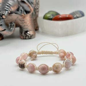 May include: A macramé bracelet with pink and beige gemstone beads. The bracelet is displayed on a white surface, with a decorative elephant statue and a bowl of colourful stones in the background.