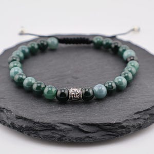 May include: A beaded bracelet featuring green and black round beads. A silver-colored cylindrical bead with an etched design is centered on the bracelet. The bracelet is strung on a black cord with an adjustable closure.